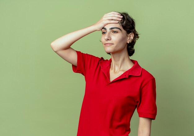 Lieta giovane ragazza caucasica con taglio di capelli pixie guardando dritto mettendo la mano sulla fronte isolata su sfondo verde oliva con spazio di copia