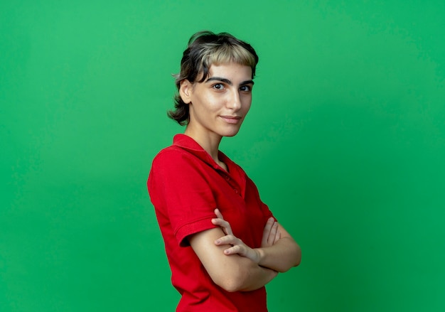 Lieta giovane ragazza caucasica con taglio di capelli pixie in piedi con la postura chiusa in vista profilo isolato su sfondo verde con spazio di copia