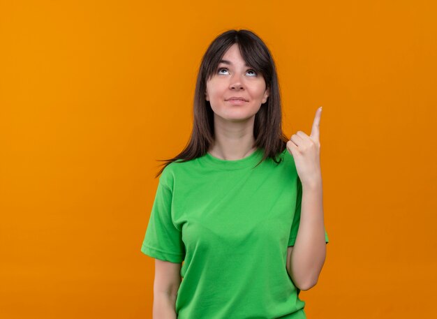 Lieta giovane ragazza caucasica in camicia verde indica e guarda su sfondo arancione isolato con spazio di copia