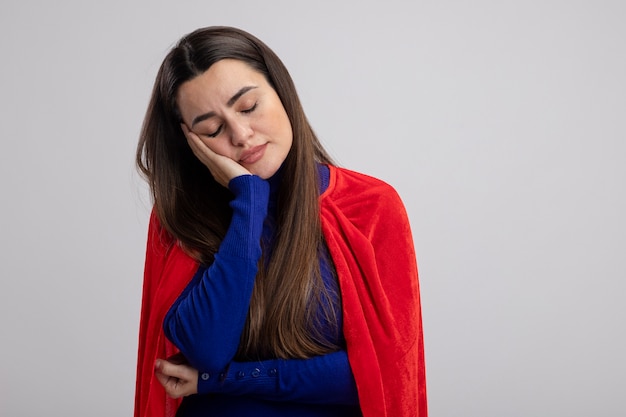 Lieta giovane ragazza del supereroe con gli occhi chiusi mettendo la mano sulla guancia isolato su sfondo bianco con copia spazio