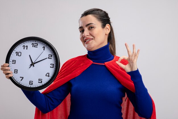 Lieta giovane ragazza del supereroe tenendo l'orologio da parete e mostrando il gesto giusto isolato su bianco