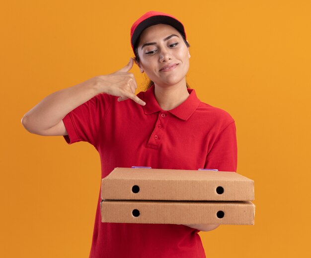 Lieta giovane ragazza delle consegne che indossa l'uniforme e il cappuccio che tiene le scatole per pizza che mostrano il gesto della telefonata isolato sulla parete arancione