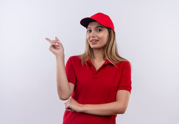 Lieta giovane ragazza delle consegne che indossa l'uniforme rossa e il cappuccio indica il lato isolato sul muro bianco con lo spazio della copia