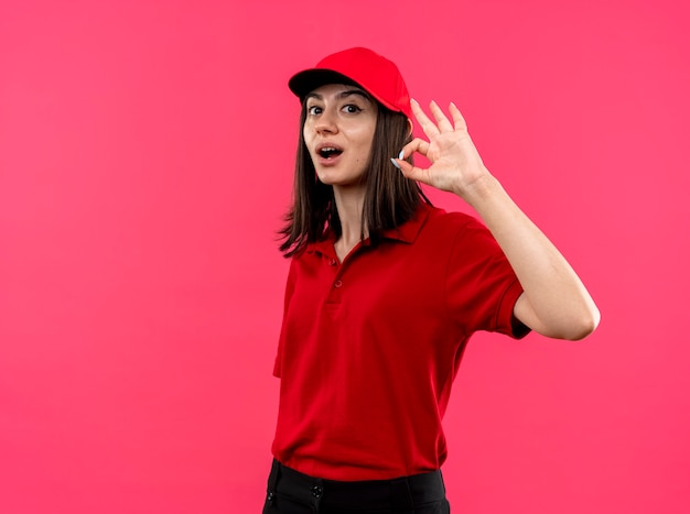 Lieta giovane ragazza delle consegne che indossa la maglietta polo rossa e cappello sorridente che mostra segno giusto in piedi sopra il muro rosa