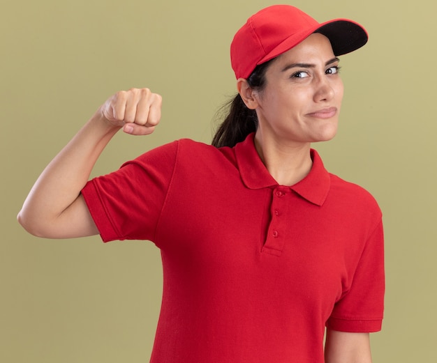 Lieta giovane ragazza di consegna che indossa l'uniforme con cappuccio che mostra un forte gesto isolato sulla parete verde oliva