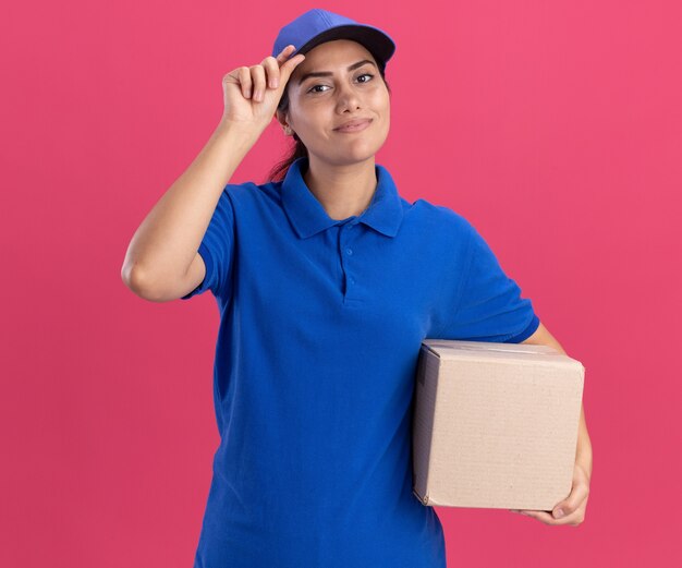 Lieta giovane ragazza di consegna che indossa l'uniforme con la scatola di contenimento del cappuccio e il cappuccio isolato sulla parete rosa
