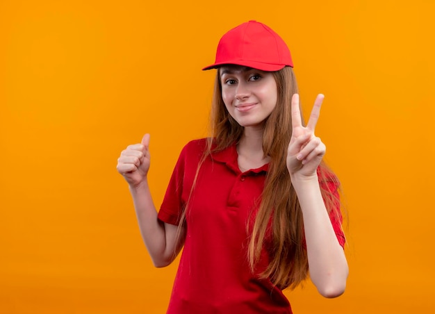 Lieta giovane ragazza di consegna in uniforme rossa che fa segno di pace e showign pollice in su sullo spazio arancione isolato con lo spazio della copia