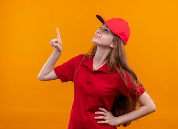 Lieta giovane ragazza di consegna in uniforme rossa che indica e che osserva in su con la mano sulla vita sullo spazio arancione isolato