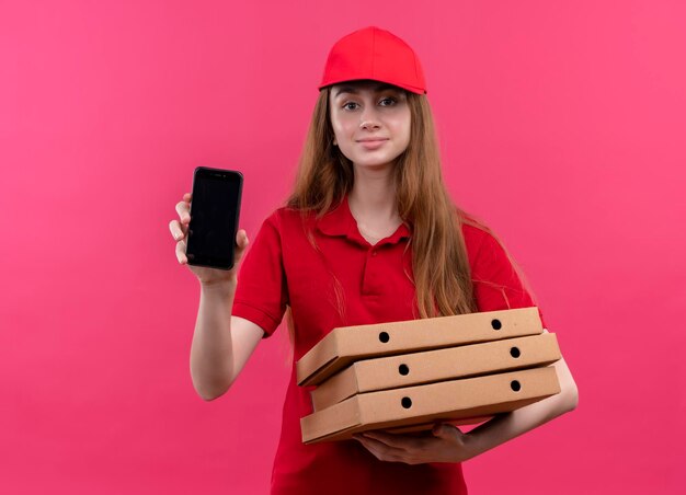 Lieta giovane ragazza di consegna in uniforme rossa che tiene i pacchetti e mostrando il telefono cellulare su uno spazio rosa isolato