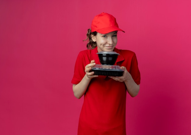Lieta giovane ragazza graziosa di consegna che indossa l'uniforme rossa e il cappuccio che tiene i contenitori per alimenti isolati su fondo cremisi con lo spazio della copia