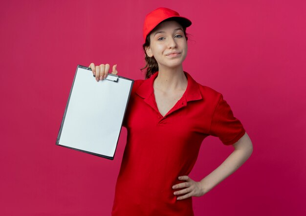 Lieta giovane ragazza graziosa di consegna in uniforme rossa e cappuccio che mostra appunti e mettendo la mano sulla vita isolata su fondo cremisi