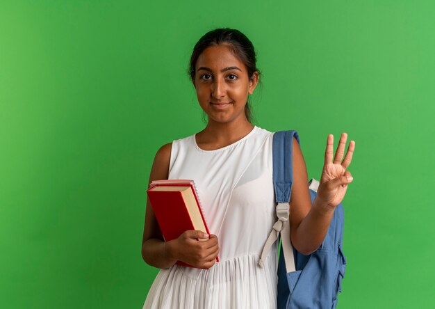 Lieta giovane studentessa che indossa la borsa indietro tenendo il libro con il taccuino e mostrando con la mano tre sul verde