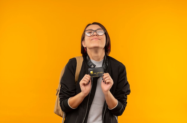 Lieta giovane studentessa con gli occhiali e borsa posteriore in possesso di carta di credito con gli occhi chiusi isolati sulla parete arancione