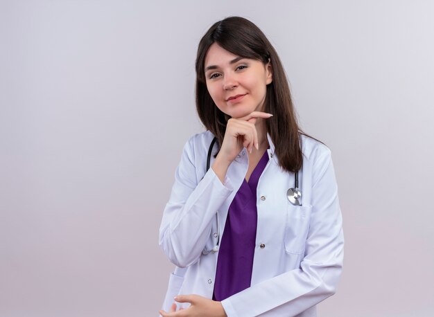 Lieta giovani donne medico in abito medico con uno stetoscopio mette la mano sul mento e guarda la telecamera su sfondo bianco isolato con spazio di copia