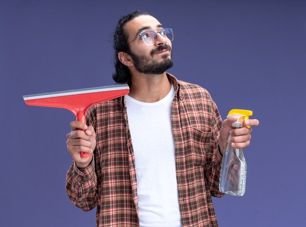 Lieta guardando il giovane bel ragazzo delle pulizie di lato che indossa la maglietta che tiene la testa del mop con il flacone spray isolato sulla parete blu