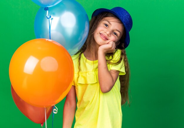 lieta piccola ragazza caucasica con cappello da festa blu mettendo la mano sul viso e tenendo palloncini di elio isolati sulla parete verde con spazio di copia