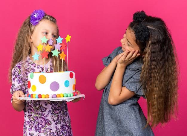 Lieta piccola ragazza caucasica guardando piccola ragazza bionda con torta di compleanno isolata sulla parete rosa con spazio copia
