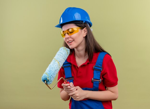 Lieta ragazza giovane costruttore con casco di sicurezza blu e con occhiali di sicurezza tiene il rullo di vernice con entrambe le mani su sfondo verde isolato