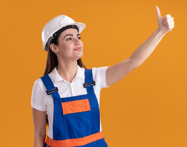 Lieto di guardare di lato la giovane donna del costruttore in uniforme che mostra il pollice in alto isolato sul muro arancione