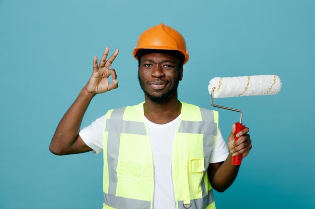 Lieto di mostrare gesto ok giovane costruttore afroamericano in uniforme con spazzola a rullo isolata su sfondo blu