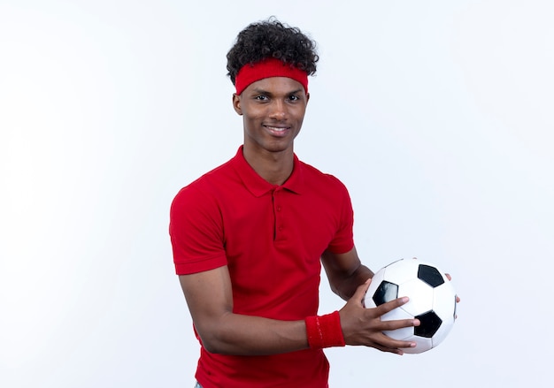 Lieto giovane afro-americano sportivo uomo che indossa la fascia e il braccialetto tenendo fuori la sfera a lato isolato su sfondo bianco