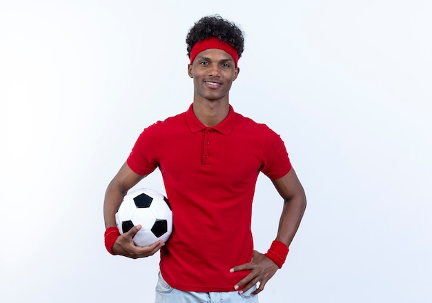 Lieto giovane afro-americano sportivo uomo che indossa la fascia e il braccialetto tenendo palla e mettendo la mano sul fianco isolato su sfondo bianco