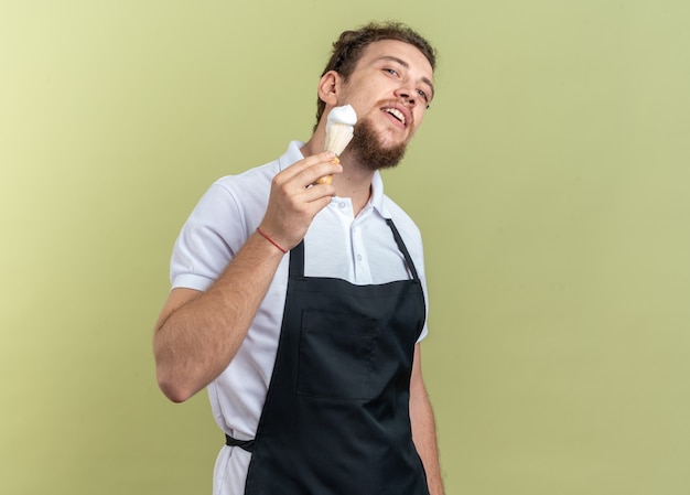 Lieto giovane barbiere maschio che indossa l'uniforme tenendo il pennello da barba isolato su parete verde oliva