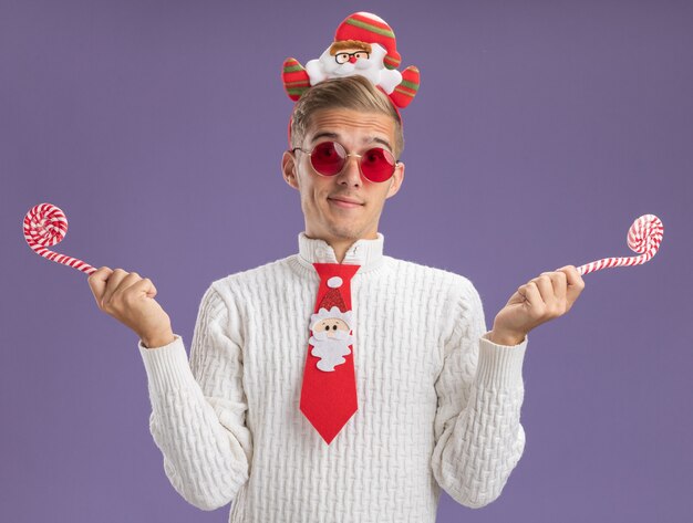 Lieto giovane bel ragazzo che indossa la fascia di Babbo Natale e cravatta con gli occhiali che tengono i bastoncini di zucchero di Natale che guarda l'obbiettivo isolato su sfondo viola