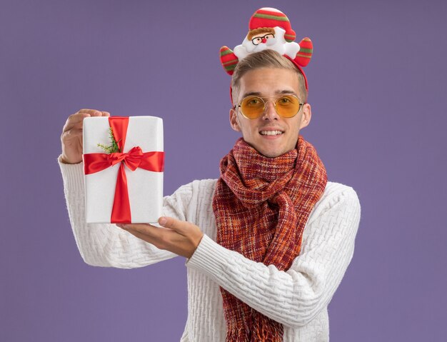 Lieto giovane bel ragazzo che indossa la fascia di Babbo Natale e sciarpa che tiene il pacchetto regalo guardando la telecamera isolata su sfondo viola