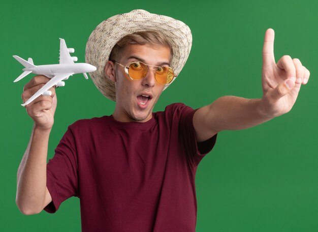 Lieto giovane bel ragazzo indossa una camicia rossa con gli occhiali e il cappello che tiene aeroplano giocattolo tendendo il dito alla macchina fotografica isolata sulla parete verde