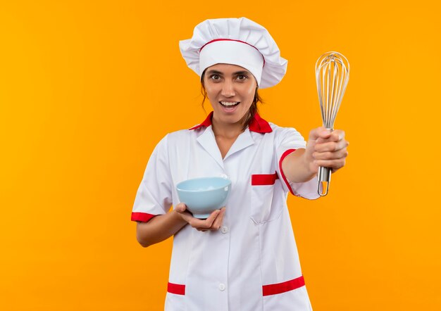 Lieto giovane cuoco femminile che indossa la ciotola uniforme della holding dello chef tenendo fuori la frusta sulla parete gialla isolata con lo spazio della copia