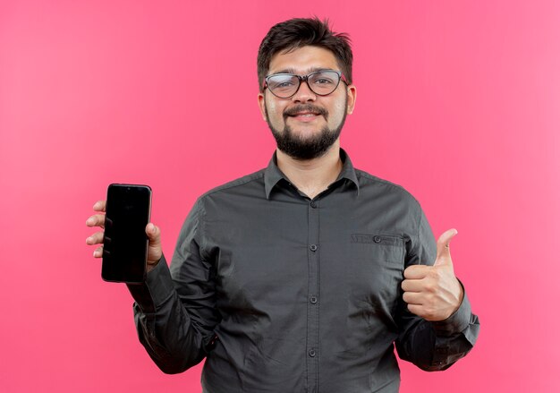 Lieto giovane imprenditore con gli occhiali tenendo il telefono con il pollice in alto isolato sul muro rosa