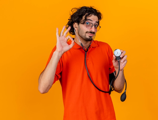 Lieto giovane uomo malato caucasico con gli occhiali e lo stetoscopio che tiene lo sfigmomanometro facendo segno giusto isolato sulla parete arancione con lo spazio della copia