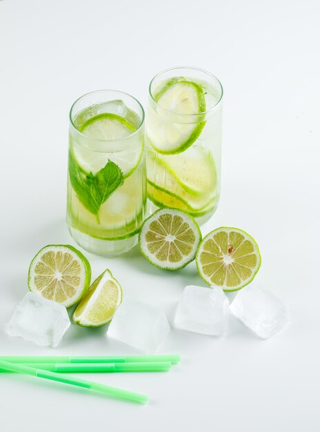 Limonata con limoni, cannucce, basilico, cubetti di ghiaccio in bicchieri su bianco, veduta dall'alto.