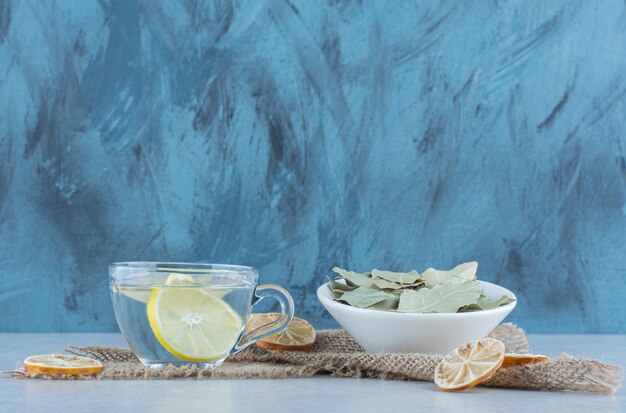 Limonata e limone secco affettato accanto a una ciotola di foglie su un asciugamano su marmo.