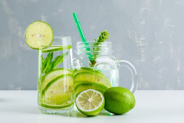 Limonata in vetro e barattolo di vetro con limone, erbe, paglia vista laterale su bianco e gesso