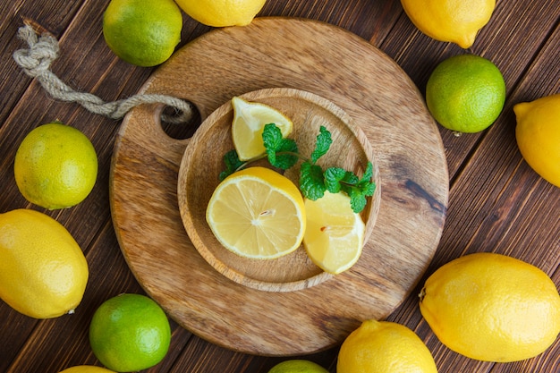 Limoni con lime, foglie in un piatto di legno su legno e tagliere, piatto disteso.