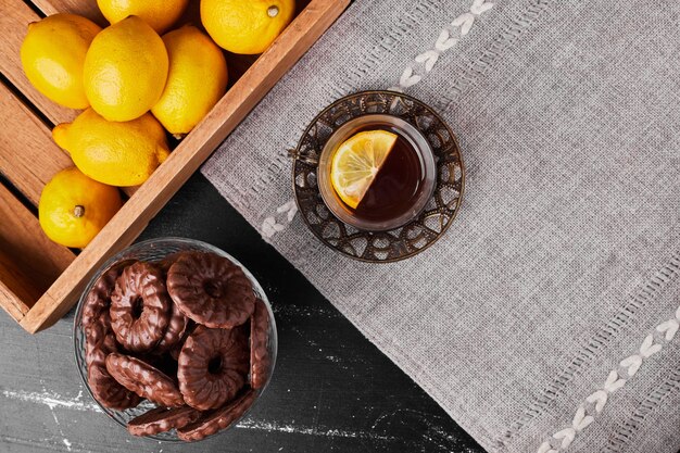Limoni isolati su uno sfondo nero in un vassoio di legno con biscotti e un bicchiere di tè intorno.
