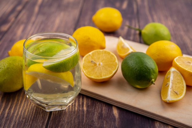 Limoni vista laterale con limette su un tagliere con un bicchiere di acqua detox su uno sfondo di legno