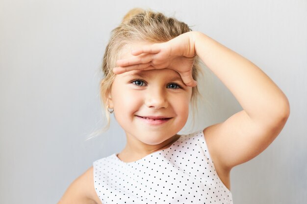 Linguaggio del corpo. Inquadratura orizzontale di carina bambina allegra con i capelli biondi raccolti tenendo il palmo sulla fronte come se guardasse lontano, cercando di vedere qualcosa di lontano, sorridendo felice