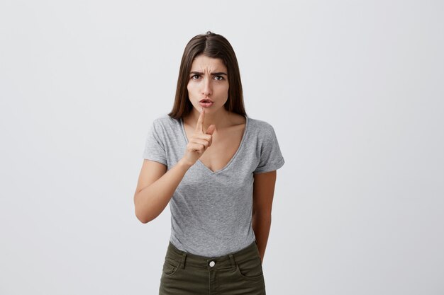 Linguaggio del corpo. Madre che mostra al suo bambino di non urlare in pizzo pubblico. Giovane bella donna seria con capelli lunghi scuri in abbigliamento casual che tiene il dito indice davanti alla bocca