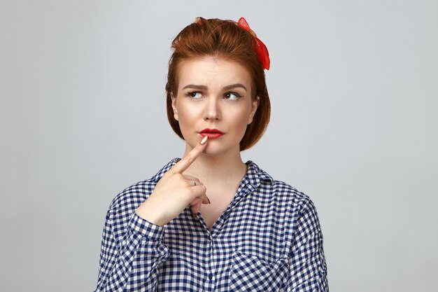 Linguaggio del corpo. Ragazza carina incerta premurosa che indossa acconciatura vintage e trucco luminoso tenendo il dito sulle labbra e guardando di traverso, sentendosi dubbioso su qualcosa, posa in studio