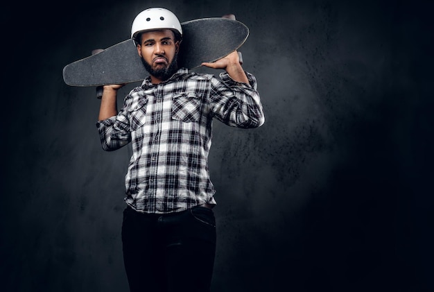 Lo skater nero vestito con una maglietta in pile tiene una tavola lunga su sfondo grigio in uno studio.