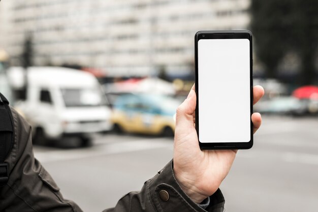 Lo smart phone della tenuta della mano di una persona che mostra lo schermo in bianco bianco contro la strada vaga