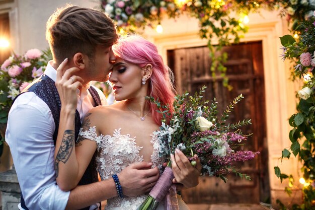 Lo sposo alla moda abbraccia da dietro bella sposa con i capelli rosa