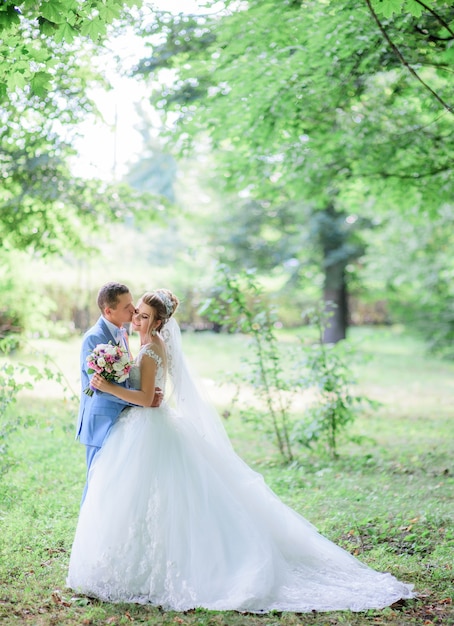 Lo sposo baci tenera guancia della sposa in piedi con lei nel parco verde