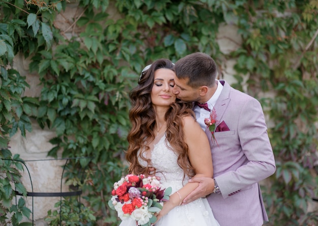 Lo sposo bello sta baciando la bella sposa all'aperto vestita in abbigliamento da sposa alla moda