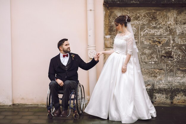 Lo sposo sulla sedia a rotelle tiene la mano della sposa in piedi davanti alla vecchia casa sulla strada