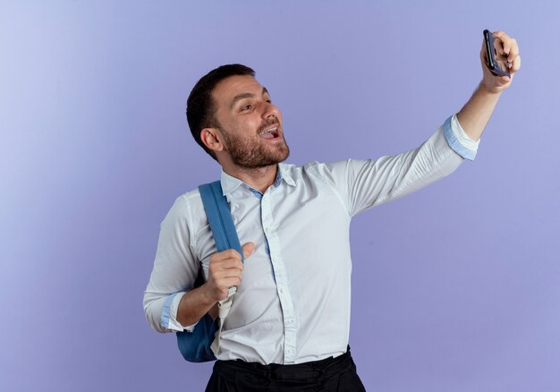 Lo zaino da portare dell'uomo bello allegro tiene il telefono che prende selfie isolato sulla parete viola