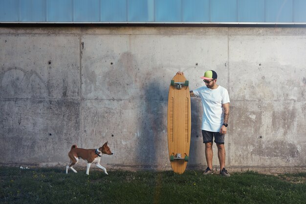 Longboarder barbuto e tatuato in piedi accanto a un muro di cemento guardando un cane basenji marrone e bianco in avvicinamento